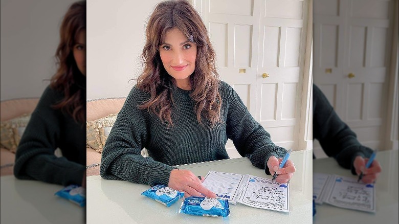 Idina Menzel sitting at a desk and writing in a book with Rice Krispie Treats