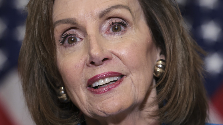 Nancy Pelosi speaking in front of American flag with slight smile