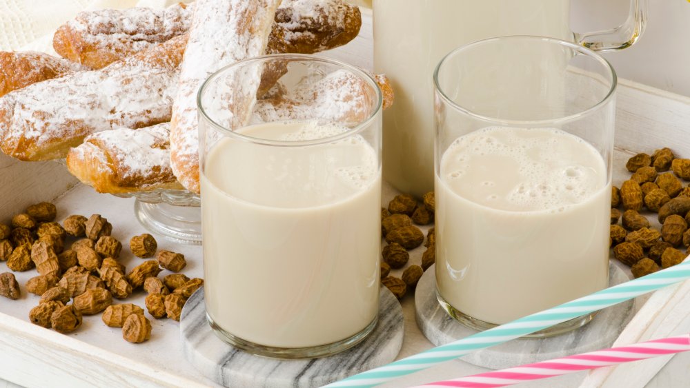 An image of horchata de chufa. The glass and pitcher containing the beverage are surrounded by chufas, or "tiger nuts."