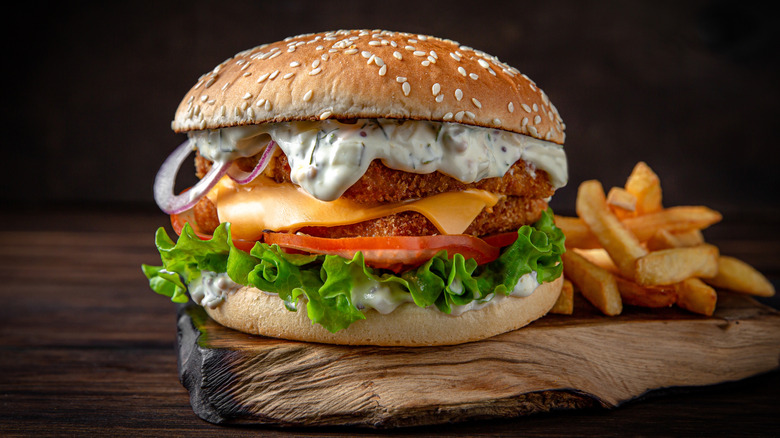 A fish sandwich with tartar sauce, onions, cheese, tomato, and lettuce beside fries