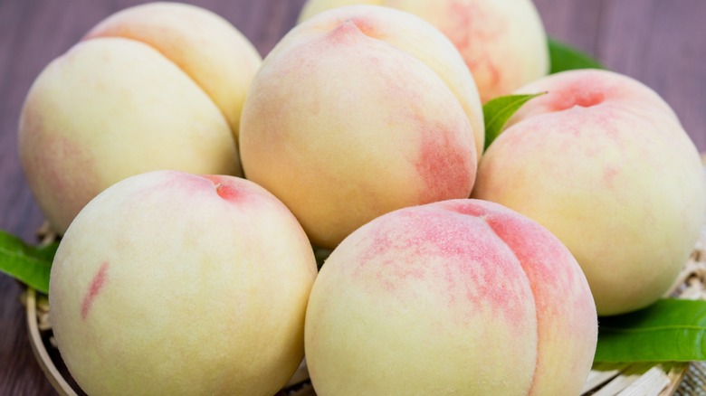 Five white peaches sitting on a wicker plate on a wooden table