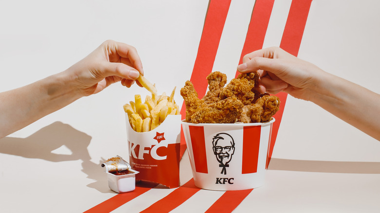 two hands picking food from KFC containers of french fries and fried chicken on white background with red diagonal stripes