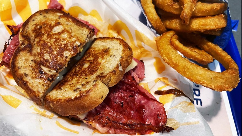 A crispy sandwich leaking meat, cheese, and sauerkraut into a basket. Some onion rings sit on the side.