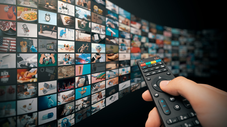 A hand holds up a TV remote to a wall of screens.