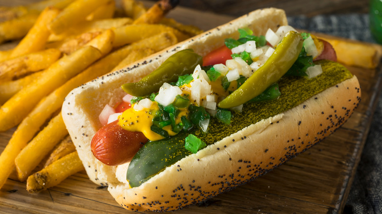 Chicago style hot dog with pickle, sport peppers, relish, onions, tomato, mustard, and celery salt on a poppy seed bun with fries on wooden cutting board