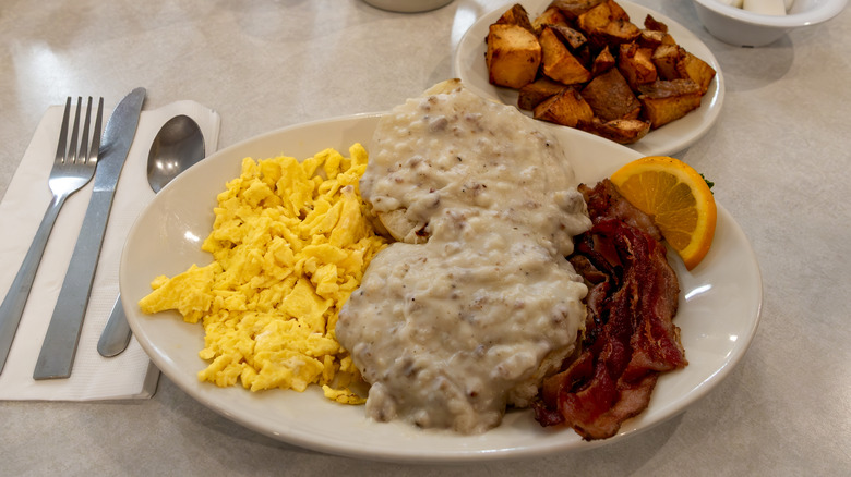 biscuits and gravy with eggs, bacon, and potatoes on a white dinner plate