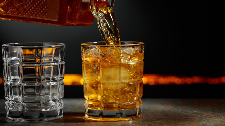 A bottle pours liquor into a glass, beside an empty glass with bright lights in the background