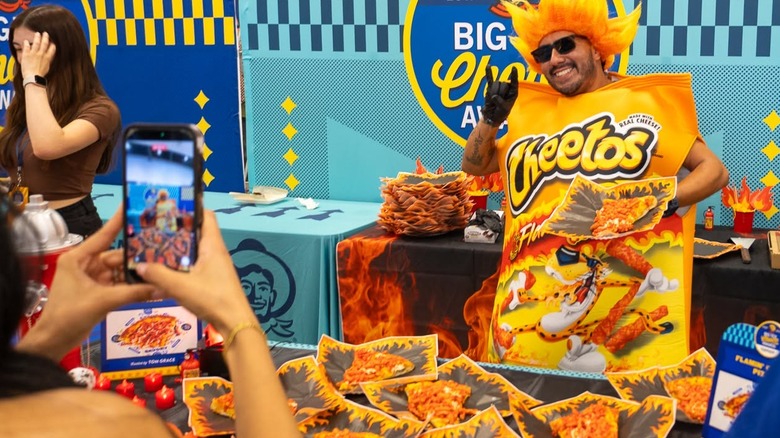 Hands taking photo on phone of Flamin' Hot Cheetos mascot with servings of Flamin' Crunch Pizza slices