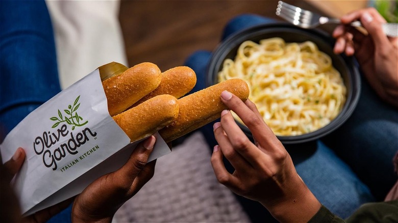 person holding Olive Garden breadsticks and takeout container of pasta