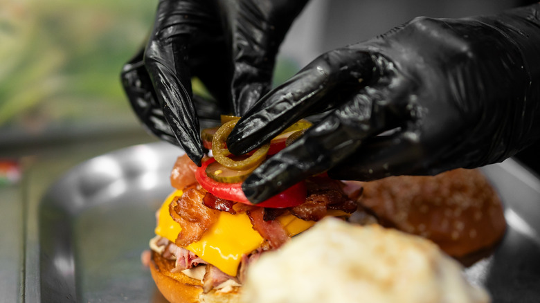 Gloved hands put jalapeños on a bacon cheeseburger