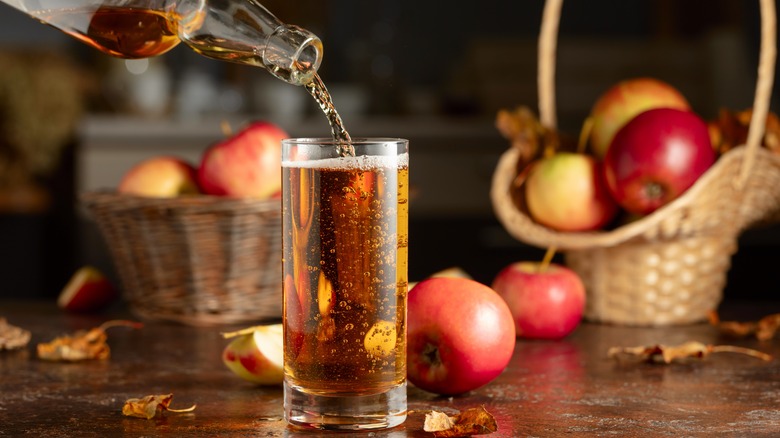 person pouring apple cider in glass with baskets of apples in background