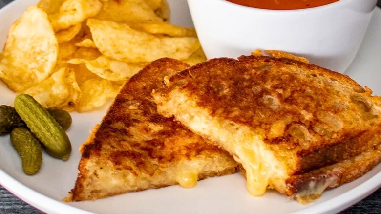 A plate with grilled cheese, tomato soup, pickles and chips