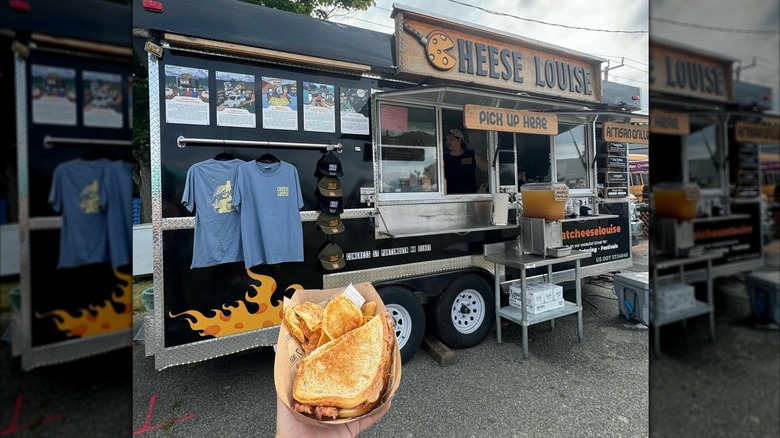 Hand holding a grilled cheese, with a food truck on the background