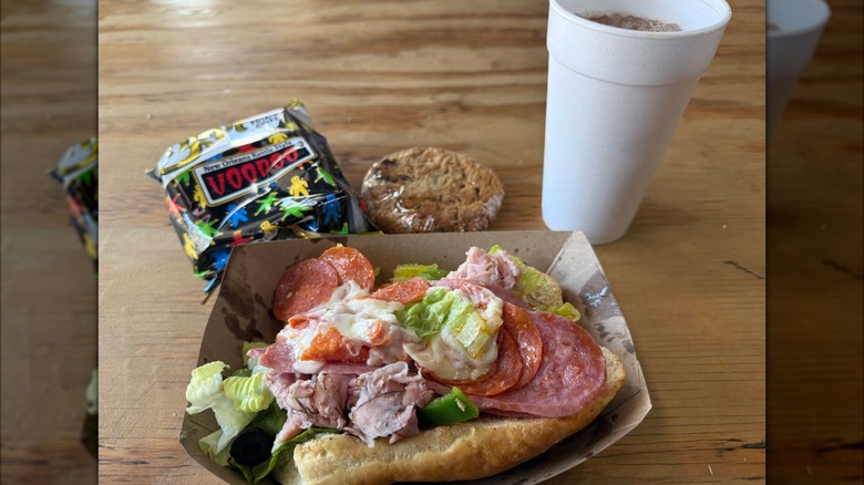 An open-faced Italian sub on a roll piled with deli meats, topped with shredded lettuce and peppers, served in a tray with a bag of Voodoo chips, a wrapped cookie, and a foam cup