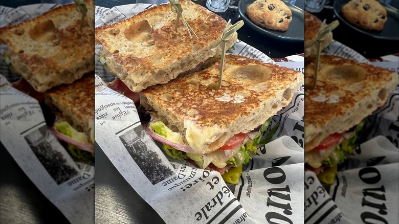 An Italian sandwich cut in half, layered with deli meats, lettuce, tomato, and onions, served on deli paper with a cookie on the side