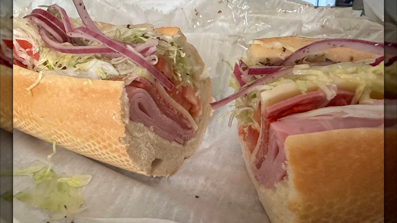 A halved Italian sub on a roll filled with layers of deli meats, cheese, shredded lettuce, tomato, and thinly sliced red onions