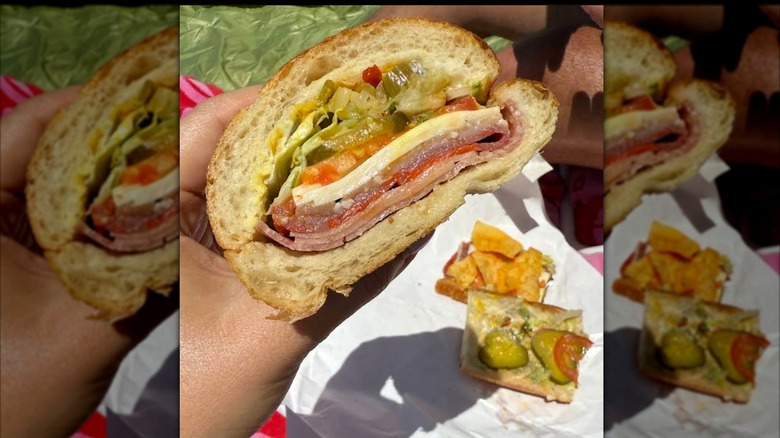 A hand holding half of an Italian sub filled with deli meats, provolone, shredded lettuce, tomatoes, and chopped pickles, with chips and the other half of the sandwich in the background