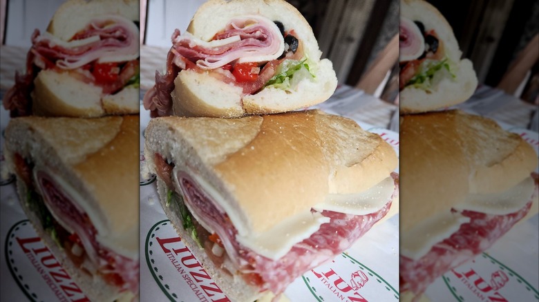 Two halves of an Italian sub with layers of salami, ham, provolone, tomatoes, lettuce, and black olives on a soft roll