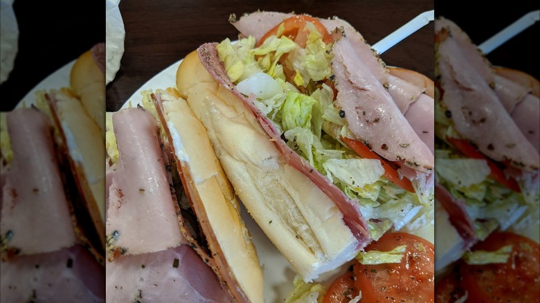 A plated Italian sub cut into halves, packed with slices of turkey, ham, salami, provolone, shredded lettuce, and tomatoes, seasoned with herbs and black pepper