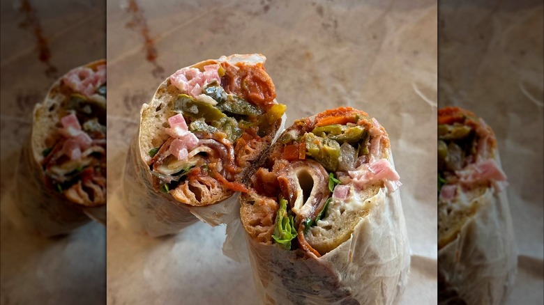 A cut Italian sandwich wrapped in paper, showing a packed cross-section of cured meats, cheese, pickled vegetables, and dressing soaked into the bread