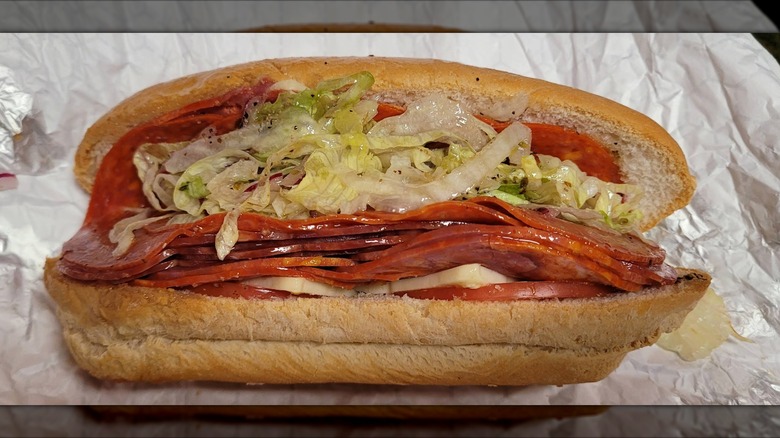 A loaded Italian sub on a long roll stacked with layers of pepperoni, salami, provolone, tomatoes, shredded lettuce, and oil