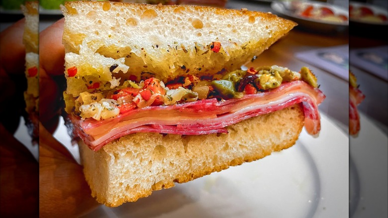 A close-up of an Italian sandwich on airy focaccia filled with layers of salami, ham, and provolone, topped with chopped pickled vegetables and herbs