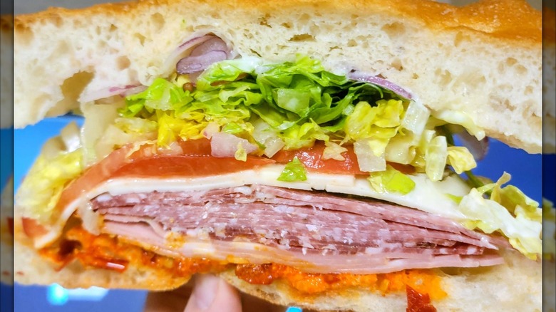 A cross-section of an Italian sub packed with layers of deli meats, provolone, shredded lettuce, tomato, and red pepper spread