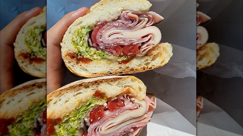 Two halves of an Italian sub held in hand, showing thick layers of ham, salami, provolone, shredded lettuce, tomato, and dressing