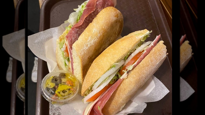 Two Italian sub halves filled with deli meats, cheese, lettuce, tomato, and onions, served with a side of pickled vegetables