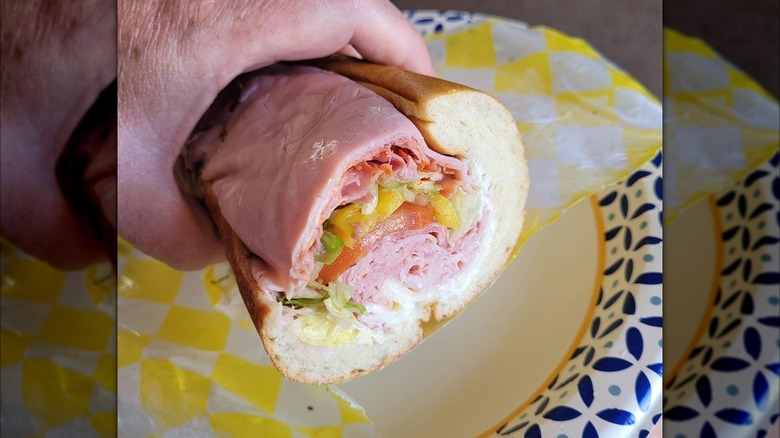 A close-up of an Italian sub held in a hand, loaded  with layers of deli meats and cheese, lettuce, tomato, and banana peppers