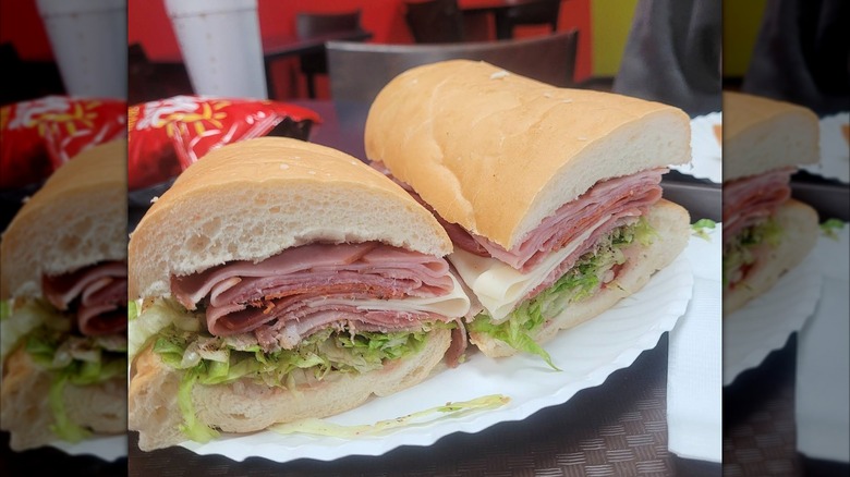 Two halves of an Italian sub on a soft roll, stacked with layers of deli meats, cheese, and lettuce, served on a white paper plate