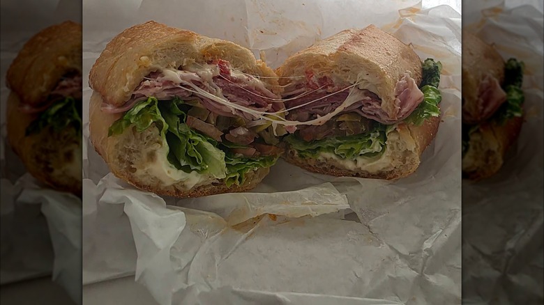 A halved Italian sub on a crusty roll, layered with deli meats, lettuce, chopped vegetables, and melted cheese on deli paper