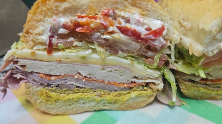 A close-up of a halved Italian sub stacked with layers of deli meats, cheese, lettuce, tomato, and mayo and mustard as condiments