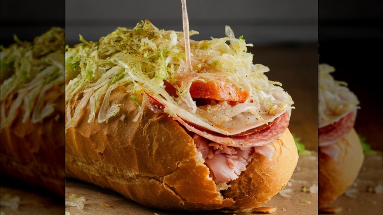 A crusty Italian sub piled high deli meats and cheese, tomato, and finely shredded lettuce, with a toothpick holding it together