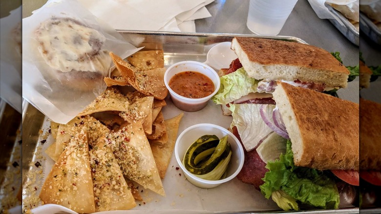 A deli tray with an Italian sub filled with meats, cheese, lettuce, and tomato on toasted bread, served alongside chips, dipping sauces, pickles, and a cinnamon roll