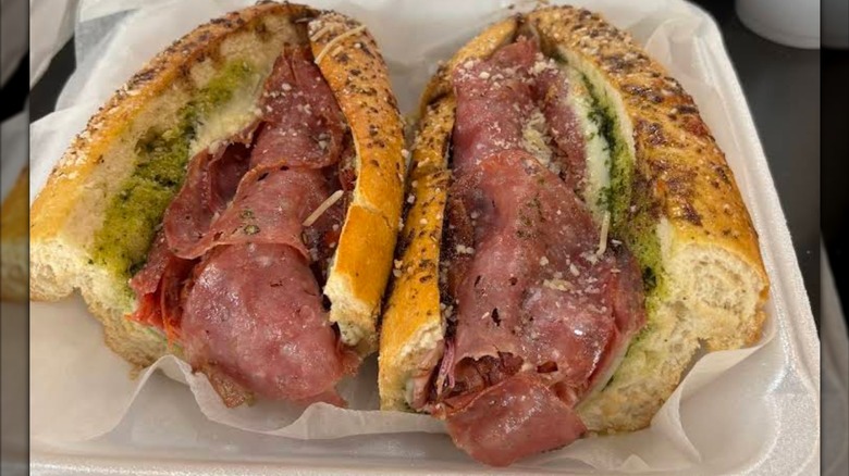 A toasted Italian sub in a takeout container, layered with deli meats, melted cheese, and pesto