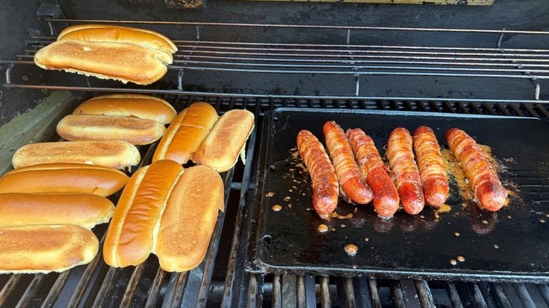 Hot dog buns and bacon-wrapped hot dogs cooking on grill