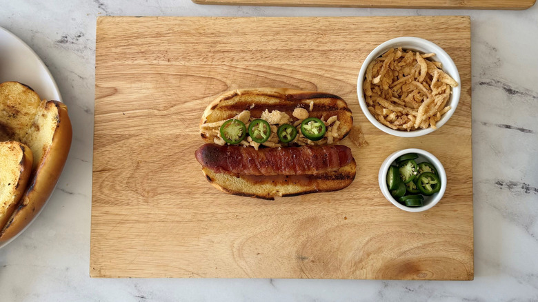 Bacon-wrapped hot dog in bun topped with jalapeño slices and crispy onions