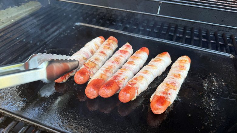 Tongs turning bacon-wrapped hot dog on grill skillet