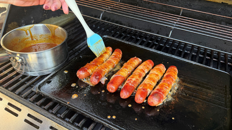 Brushing bacon-wrapped hot dogs with sauce on grill