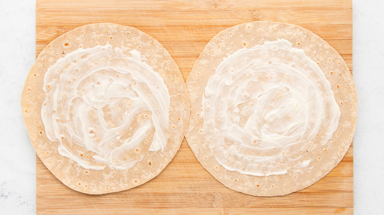 Two flour tortillas spread with mayonnaise on wooden cutting board