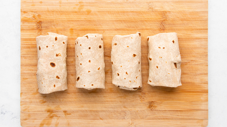 Four filled and rolled flour tortilla wraps on wooden cutting board