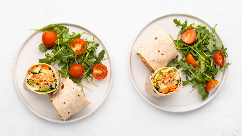 Two BBQ chicken wraps on plates with arugula and tomato salad