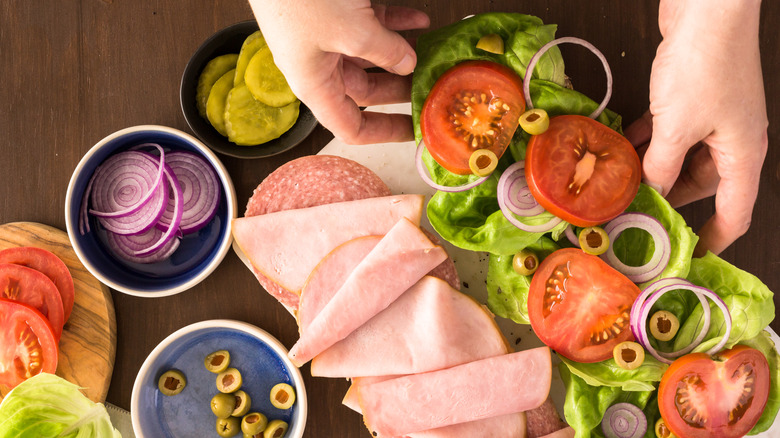 A close-up of two hands assembling a sub with bowls of chopped vegetables on the table