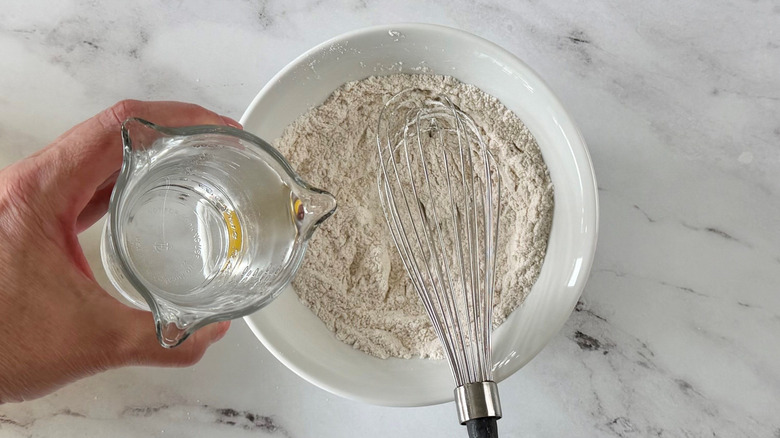 Hand pouring water into flour mixture in bowl with whisk