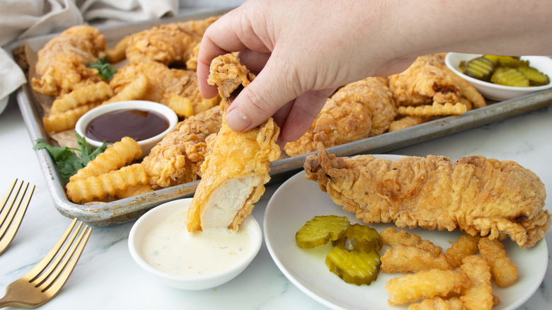 hand dipping chicken tenders in sauce next to baking tray of tenders and fries