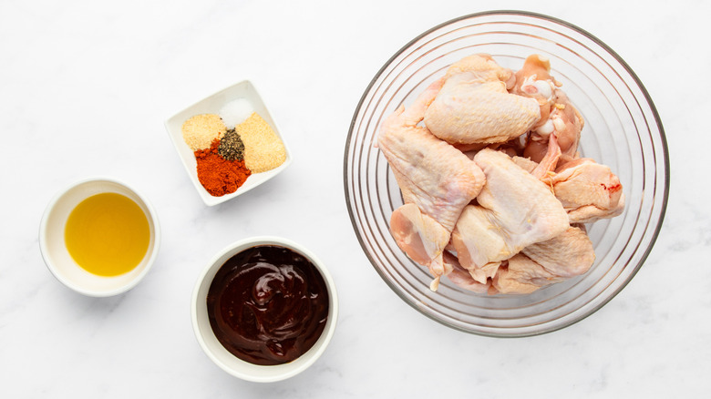 Raw chicken wings in a bowl next to seasonings, bowl of barbecue sauce, and olive oil