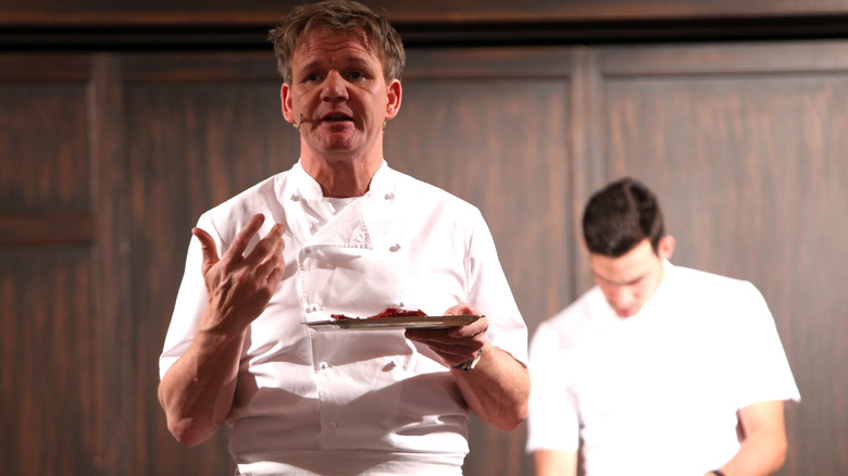 Gordon Ramsay holding a plate and explaining while conducting a cooking lesson in front of another chef in background