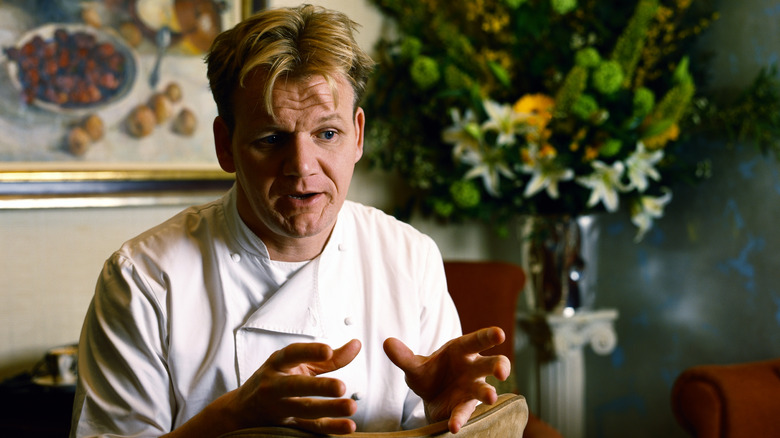 a young Gordon Ramsay explaining and gesturing with hands in front of decorative painting and flowers