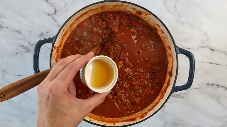 Hand holding small bowl of bourbon over chili in pot